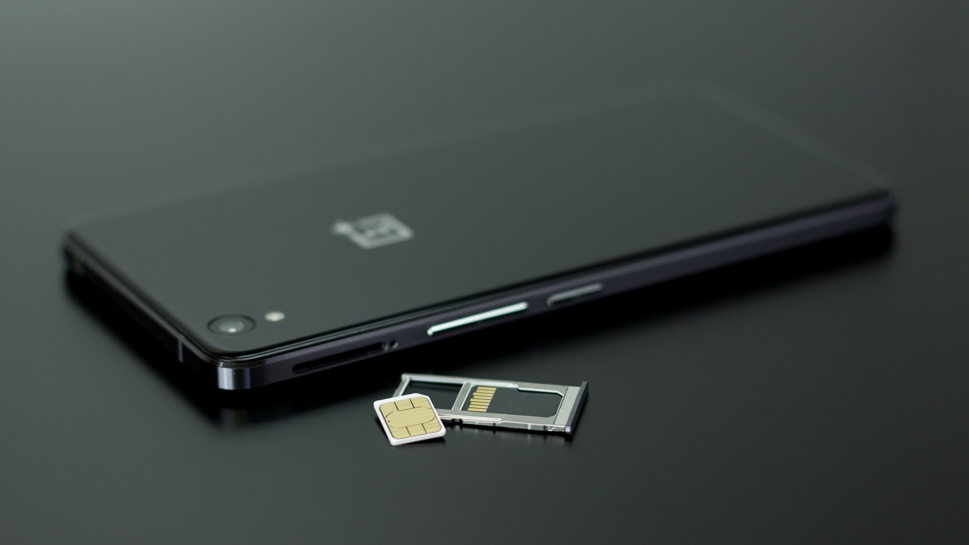 Fekete okostelefon hátulról, mellette egy kivett SIM-kártya tálca látható sötét háttéren. A tálcában egy nano-SIM kártya és egy microSD kártya található. A telefonon egy kamera és egy vaku van a bal felső sarokban, középen pedig egy logó látható. A kép a készülék beállítását vagy kártyacseréjét illusztrálja.