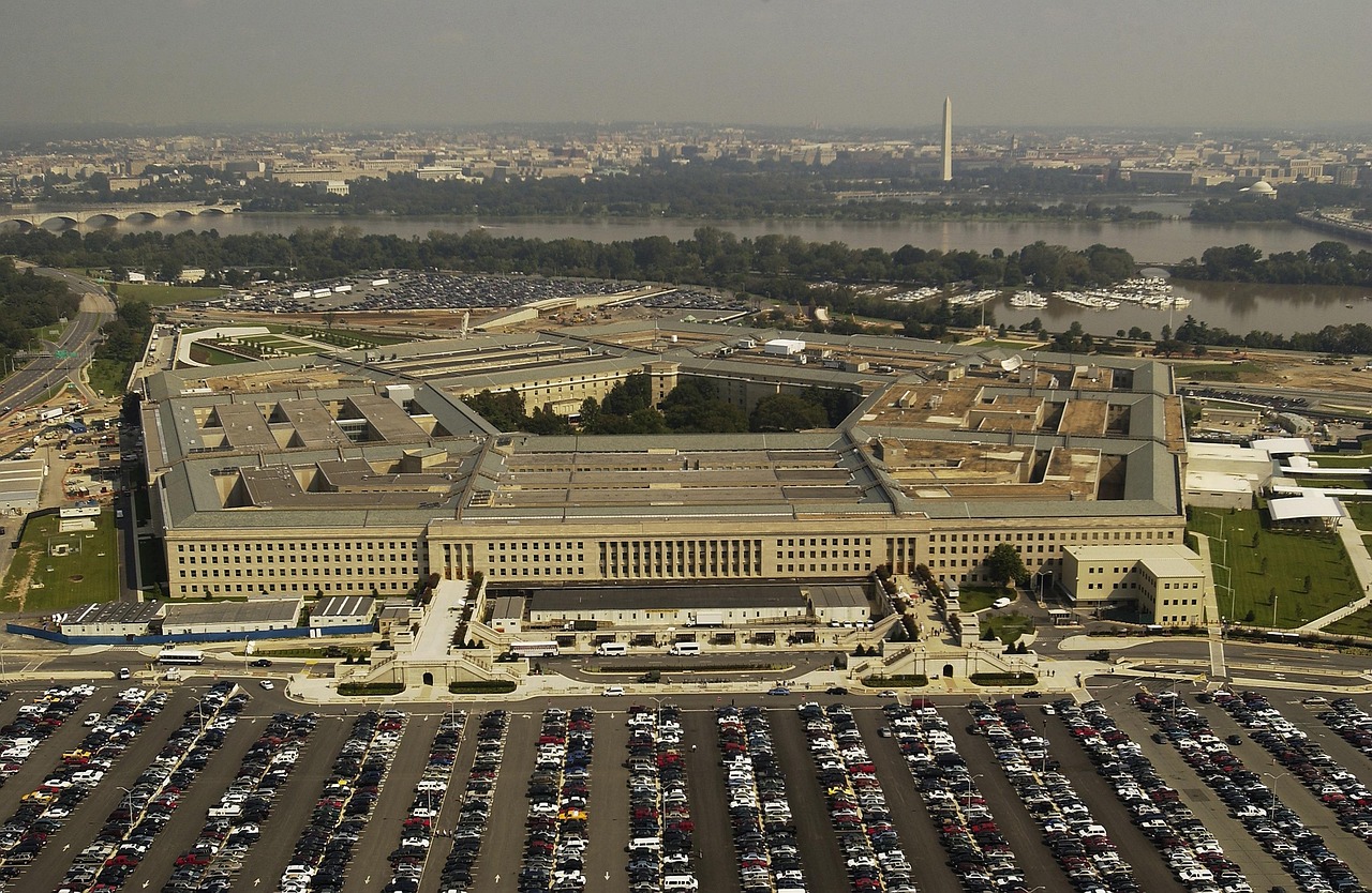 A Pentagon légifotója, egy ötszög alakú, masszív épület Washington D.C. közelében, körülötte hatalmas parkolókkal, zöld területekkel és a Potomac folyóval a háttérben, a város látképével és a Washington-emlékművel a távolban.