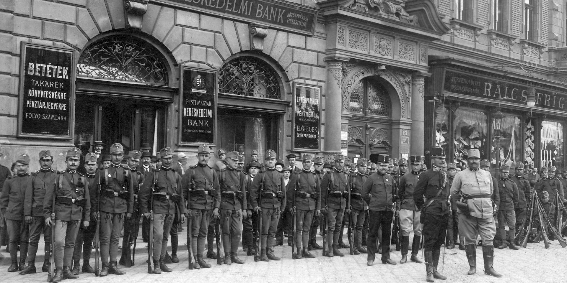 Fekete-fehér történelmi fotó számos magyar katonáról, akik egy bank épülete előtt sorakoznak az utcán, korabeli egyenruhában és fegyverzettel.